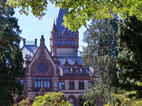 Majestic vertical shot of fairytalesque Schloss Drachenburg castle framed by green tree leaves against a clear blue sky, iconic neogothic architectural landmark and tourist destination in Koenigswin .