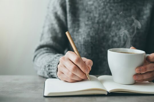 Person writing in a notebook while holding a steaming cup of coffee