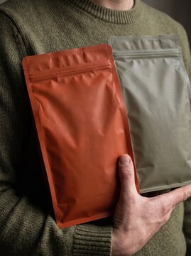 Person holding two blank stand-up pouches, one orange and one olive green, against a plain background.