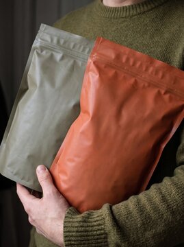 Close-up of a person holding two plain, unlabelled stand-up pouches, one olive green and one terracotta, against a dark background.