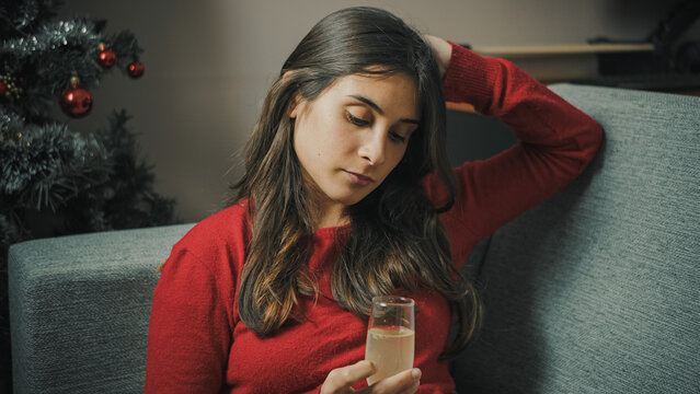 Young Hispanic woman alone at home holding a glass at Christmas with little enthusiasm, conveying subdued holiday emotions, solitude, and quiet reflection.