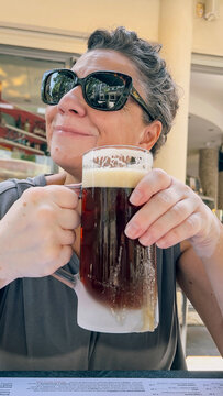A woman wearing sunglasses holds a frosted mug of dark beer while sitting at an outdoor table.