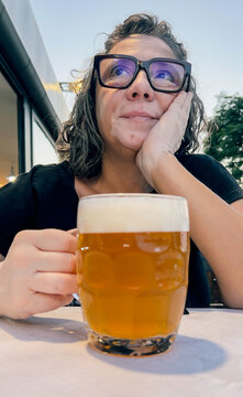 A woman wearing glasses looks away while holding a glass of beer outdoors.