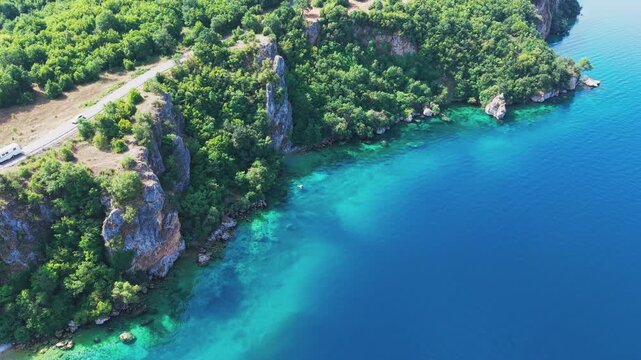Drone view of a scenic road winding along rocky, forested cliffs and the turquoise waters of Lake Ohrid near Ohrid, North Macedonia. Natural shoreline, secluded beach and clear water on a sunny summer