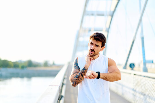 Young man monitoring heart rate with smartwatch and fingers on neck after intense workout, standing on a modern bridge