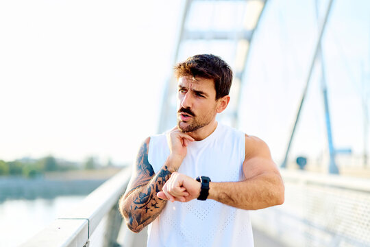 Male athlete with tattoos checking his pulse rate and fitness tracker on a bridge outdoors, monitoring health and progress