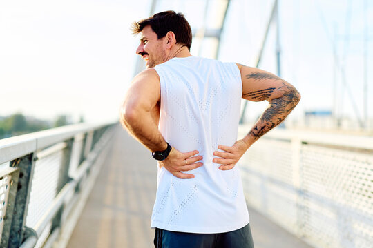 Man holding lower back, experiencing muscle strain or injury during workout on a bridge. Health problem and sport concept