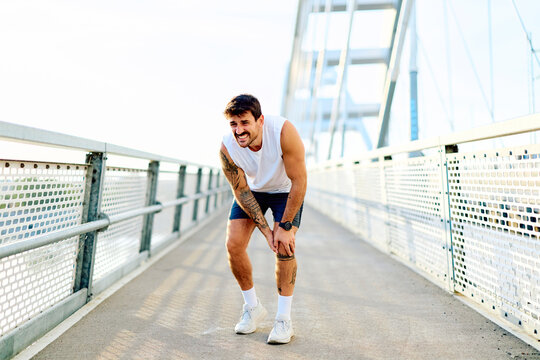 Athlete holding his injured knee after a run, suffering from a sports injury during a workout on a bridge