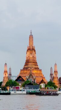 Wat Arun temple standing beside the Chao Phraya River in Bangkok, Thailand, with a traditional long-tail boat crossing the water under a bright cloudy sky, creating a scenic travel view.