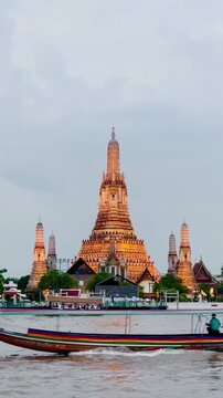 Wat Arun temple standing beside the Chao Phraya River in Bangkok, Thailand, with a traditional long-tail boat crossing the water under a bright cloudy sky, creating a scenic travel view.