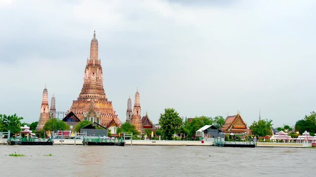 Wat Arun temple standing beside the Chao Phraya River in Bangkok, Thailand, with a traditional long-tail boat crossing the water under a bright cloudy sky, creating a scenic travel view.