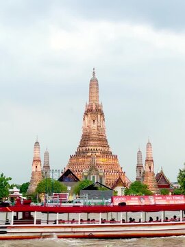 Wat Arun temple standing beside the Chao Phraya River in Bangkok, Thailand, with a traditional long-tail boat crossing the water under a bright cloudy sky, creating a scenic travel view.