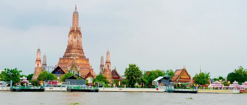 Wat Arun temple standing beside the Chao Phraya River in Bangkok, Thailand, with a traditional long-tail boat crossing the water under a bright cloudy sky, creating a scenic travel view.