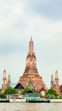 Wat Arun temple standing beside the Chao Phraya River in Bangkok, Thailand, with a traditional long-tail boat crossing the water under a bright cloudy sky, creating a scenic travel view.