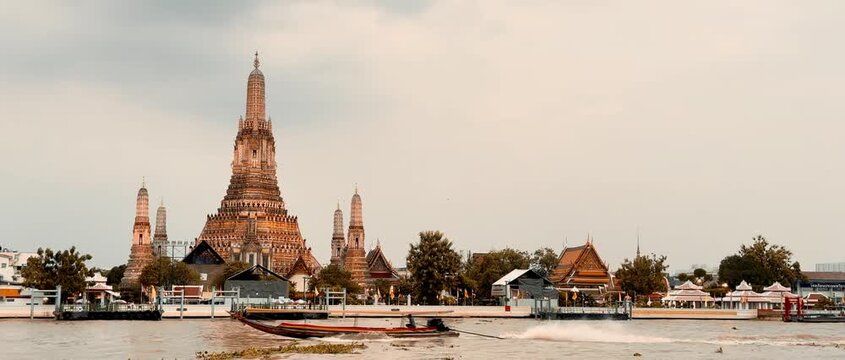Wat Arun temple standing beside the Chao Phraya River in Bangkok, Thailand, with a traditional long-tail boat crossing the water under a bright cloudy sky, creating a scenic travel view.