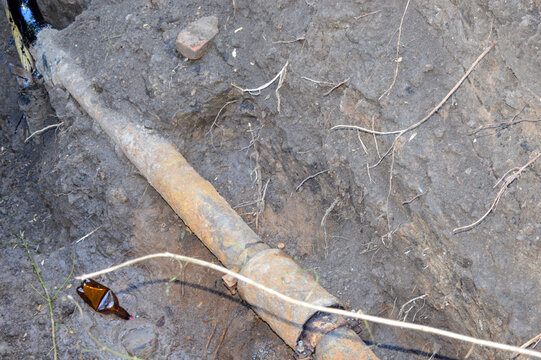 Old rusty metallic pipe lying inside an excavated soil trench, showing connection to a new black plastic section close-up. Pipeline replacement, utility repair, and infrastructure work concept.