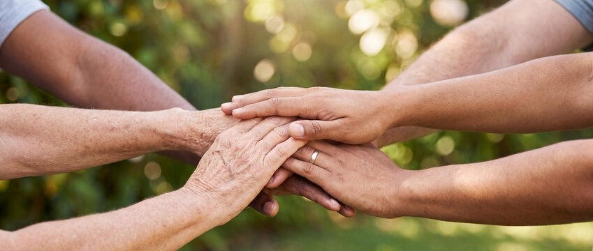 A diverse group of people in a close-up shot stack their hands together in a gesture of unity and teamwork, with a blurred green background suggesting an outdoor setting.
