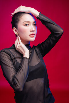 A high-fashion portrait of a woman with both arms raised elegantly, one shielding her forehead, showcasing sleek styling and striking makeup against a rich crimson background.