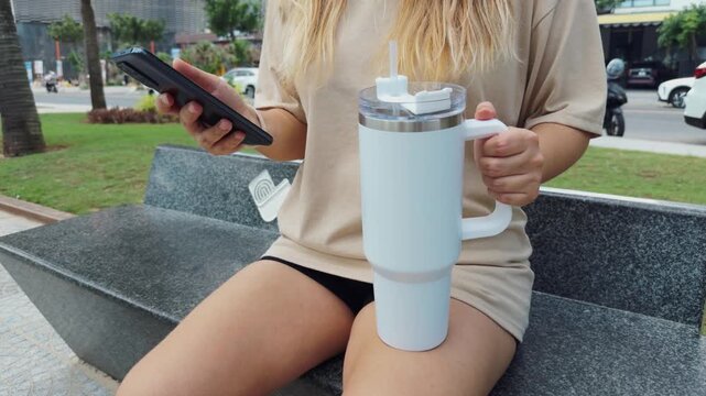Woman relaxing on a park bench, holding a white 1 liter insulated tumbler with a handle and straw, staying hydrated while using her smartphone outdoors