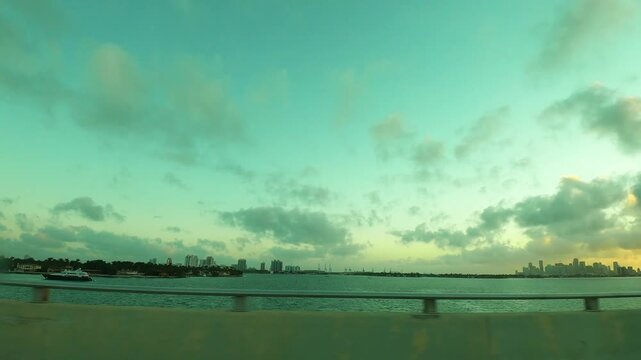 Driving along Biscayne Bay at sunset, viewed from a car window, with the Miami skyline in the background and warm evening light reflecting across the bay.