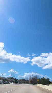 Vertical driving POV on a highway in Tampa, Florida, with a clear blue sky and white clouds, capturing the perspective of traveling by car through the American road landscape.