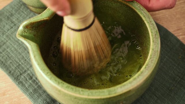 Whisking green matcha tea with traditional bamboo whisk chasen in ceramic bowl chawan to create foam. Preparing authentic Japanese matcha tea, healthy wellness beverage and detox drink.