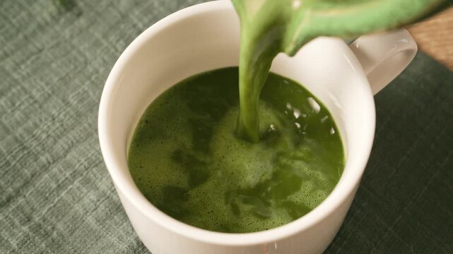 Pouring freshly prepared green matcha tea from bowl into white ceramic cup. Refreshing organic superfood matcha beverage for morning wellness routine and daily energy.