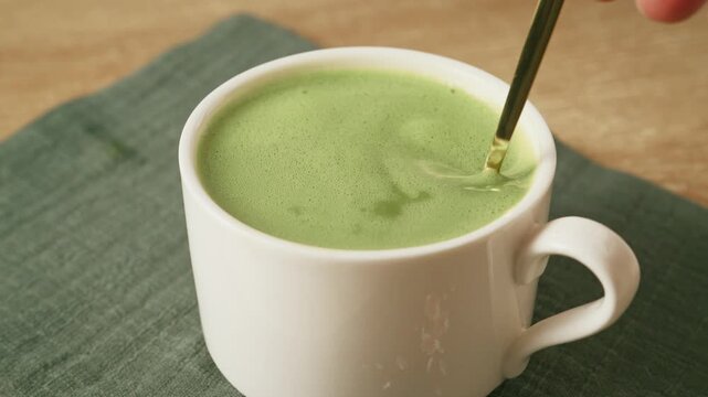 Stirring frothy green matcha latte with golden spoon in white ceramic mug. Matcha antioxidant rich detox tea for daily wellness routine.