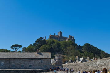 A scenic view of an island castle atop a green, tree-covered hill under a clear blue sky, with stone buildings - St Michael's Mount, Cornwall, UK © Robert