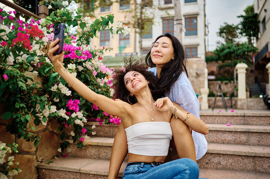 Two friends laugh and pose for a selfie on a waterfront boardwalk overlooking the sea. The smiling women enjoy a relaxed seaside outing, capturing travel memories with a smartphone against a backdrop 