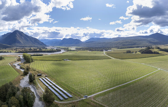 Murchison, New Zealand - 20 March 2026: Aerial view of a hop farm with rows of trellised plants, a solar panel array, and a winding river set against mountains under a cloudy sky.