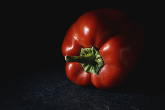 Close-up of ripe red bell pepper with vibrant green stem in moody lighting on dark background. Fresh raw vegetable for organic recipes, culinary menus, nutrition, and healthy eating.