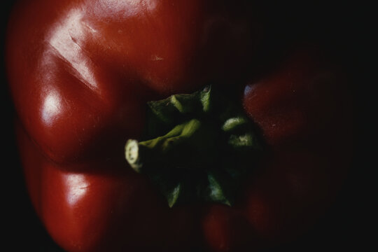 Macro shot of ripe red bell pepper with vibrant green stem in moody lighting on dark background. Fresh raw vegetable for organic recipes, culinary menus, nutrition, and healthy eating.