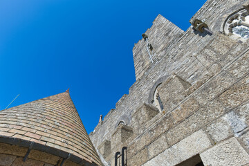 Low-angle view of a grey stone castle against a bright blue sky, featuring a conical roof and a towering wall with battlements - St Michael's Mount, Cornwall, UK © Robert