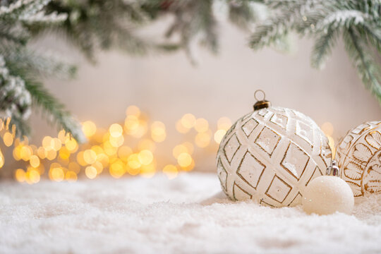 A Christmas background featuring an elegant white bauble etched with a golden diamond pattern beneath frosted pine on snow, softly lit by warm twinkling lights with generous copy space.