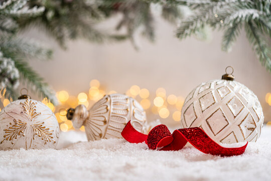 A Christmas background featuring an elegant white and gold patterned bauble with a curled red glitter ribbon beneath frosted pine on snow, softly lit by warm twinkling lights with copy space.