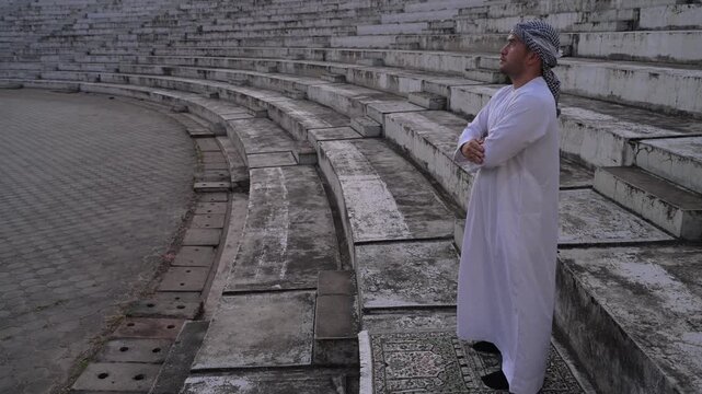 Two devoted Middle Eastern Muslim men standing on prayer mats with crossed arms performing daily religious prayers outdoors against a plain wall representing spiritual faith and traditions.