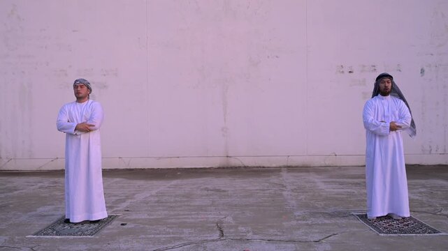 Two devoted Middle Eastern Muslim men standing on prayer mats with crossed arms performing daily religious prayers outdoors against a plain wall representing spiritual faith and traditions.