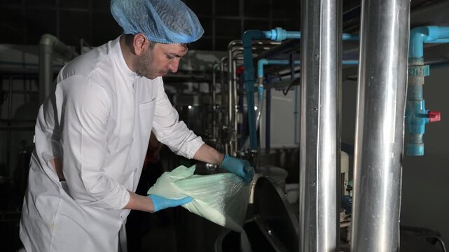 Focused professional male industrial factory worker pouring raw ingredient powder from a large bag into a stainless steel mixing tank during commercial food and beverage mass production.