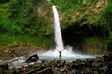 La Fortuna Waterfall cascading into a rocky pool surrounded by tropical rainforest, with a visitor highlighting the scale of this natural attraction.