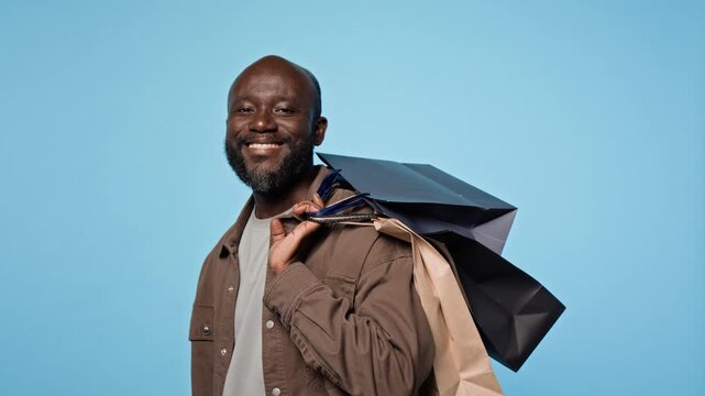 Portrait of smiling African American man putting paper shopping bags over shoulder on blue background. Clip fits retail marketing, seasonal sale promotions and consumer campaigns