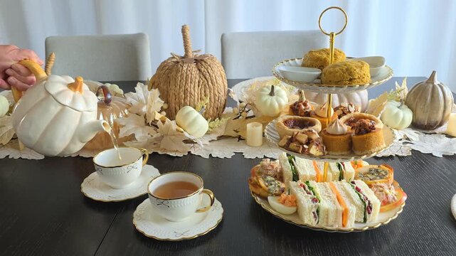 Autumn theme afternoon tea table scene with hand pouring from a pumpkin shaped teapot and a tray of scones, cozy pumpkin, pecan and apple desserts and fall theme finger sandwiches and savory items.