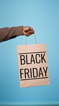Vertical video of anonymous man holding craft paper shopping bag with Black Friday sign against blue studio background. Clip suited for retail sales campaigns and holiday marketing promo