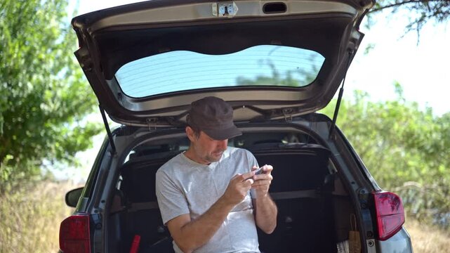 Man wearing a cap sitting in an open car trunk using a smartphone outdoors in a sunny environment. 4K.