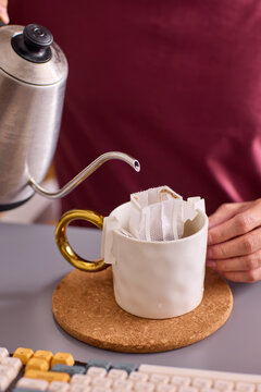 Man pouring hot water from metal kettle into clear glass mug with tea bag and ice cubes on modern living room countertop near sofa and TV under warm window light