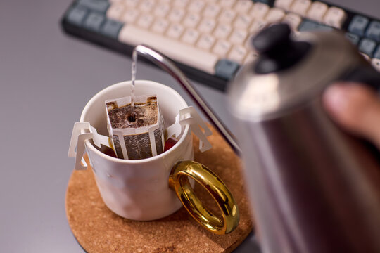 Overhead view of white ceramic coffee mug with paper pour-over dripper, placed on cork coaster beside mechanical keyboard and stainless gooseneck kettle on desk workspace