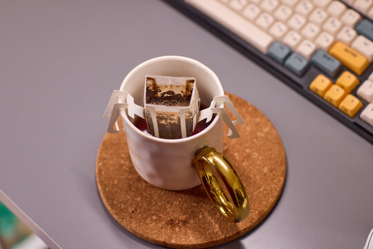 Overhead view of white ceramic coffee mug with paper pour-over dripper, placed on cork coaster beside mechanical keyboard and stainless gooseneck kettle on desk workspace