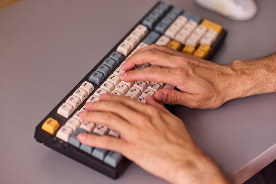 Close-up view of right-handed adult forearm and hand typing on compact mechanical keyboard with colorful keycaps on gray office desk under soft studio lighting