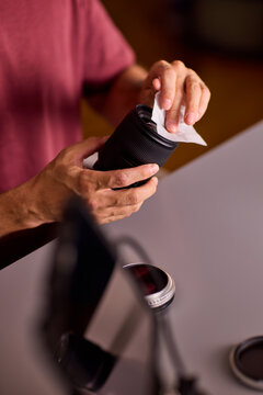 Focused young man cleaning camera lens with white microfiber cloth indoors in warm low light studio, holding lens in left hand closeup portrait side view scene