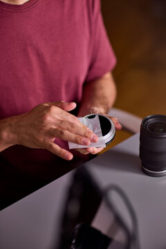 Focused young man cleaning camera lens with white microfiber cloth indoors in warm low light studio, holding lens in left hand closeup portrait side view scene